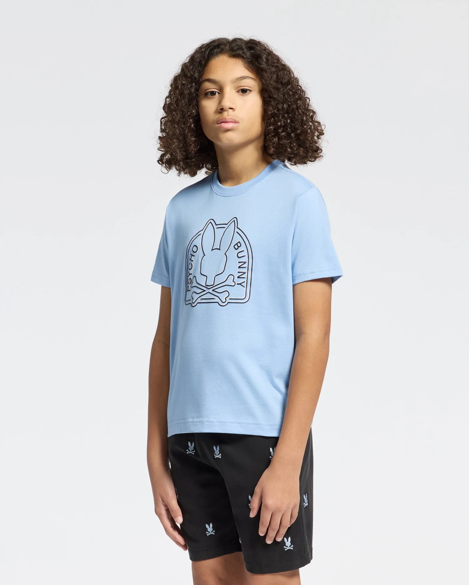 A teen is wearing the KIDS MELBOURNE GRAPHIC TEE - B0U410F200 by Psycho Bunny in light blue, made from 100% Pima cotton, with a cute bunny logo, and black shorts with small bunny designs, against a plain white background.