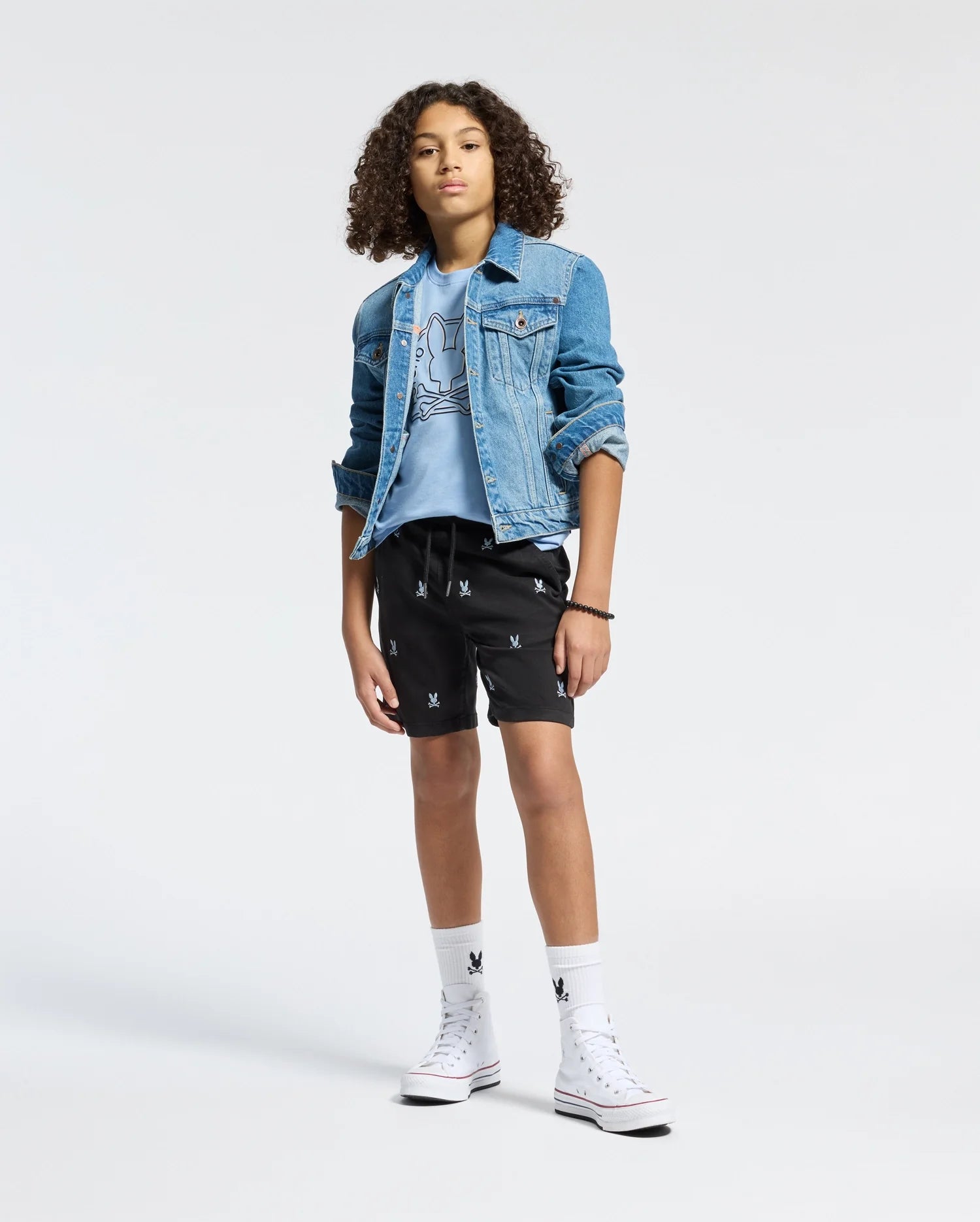 A teenager with curly hair models a Psycho Bunny KIDS MELBOURNE GRAPHIC TEE made from 100% Pima cotton. Standing on one leg in black shorts with bunny prints, they wear white socks and high-top sneakers, hands in pockets against a plain backdrop, highlighting an eco-friendly style.