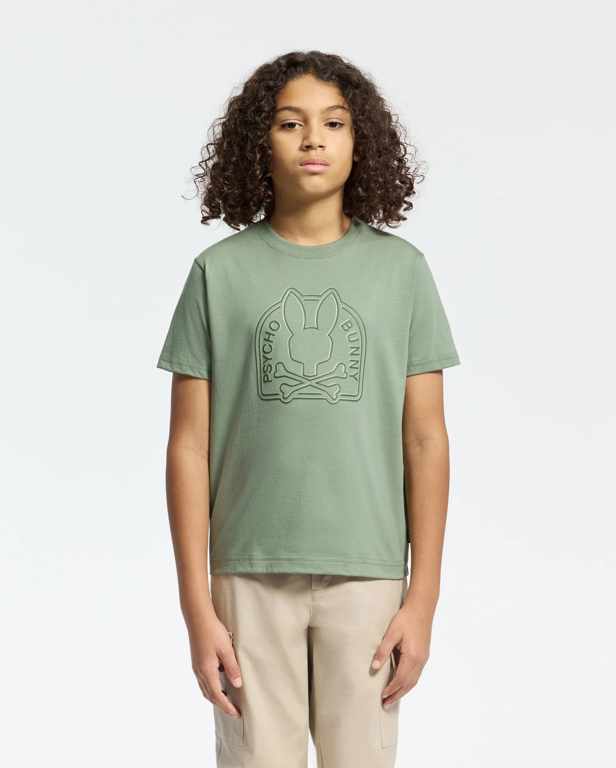 With curly hair and beige pants, a person faces forward wearing the KIDS MELBOURNE GRAPHIC TEE (B0U410F200) by Psycho Bunny, featuring a bold Bunny graphic on green Pima cotton, set against a plain white background.
