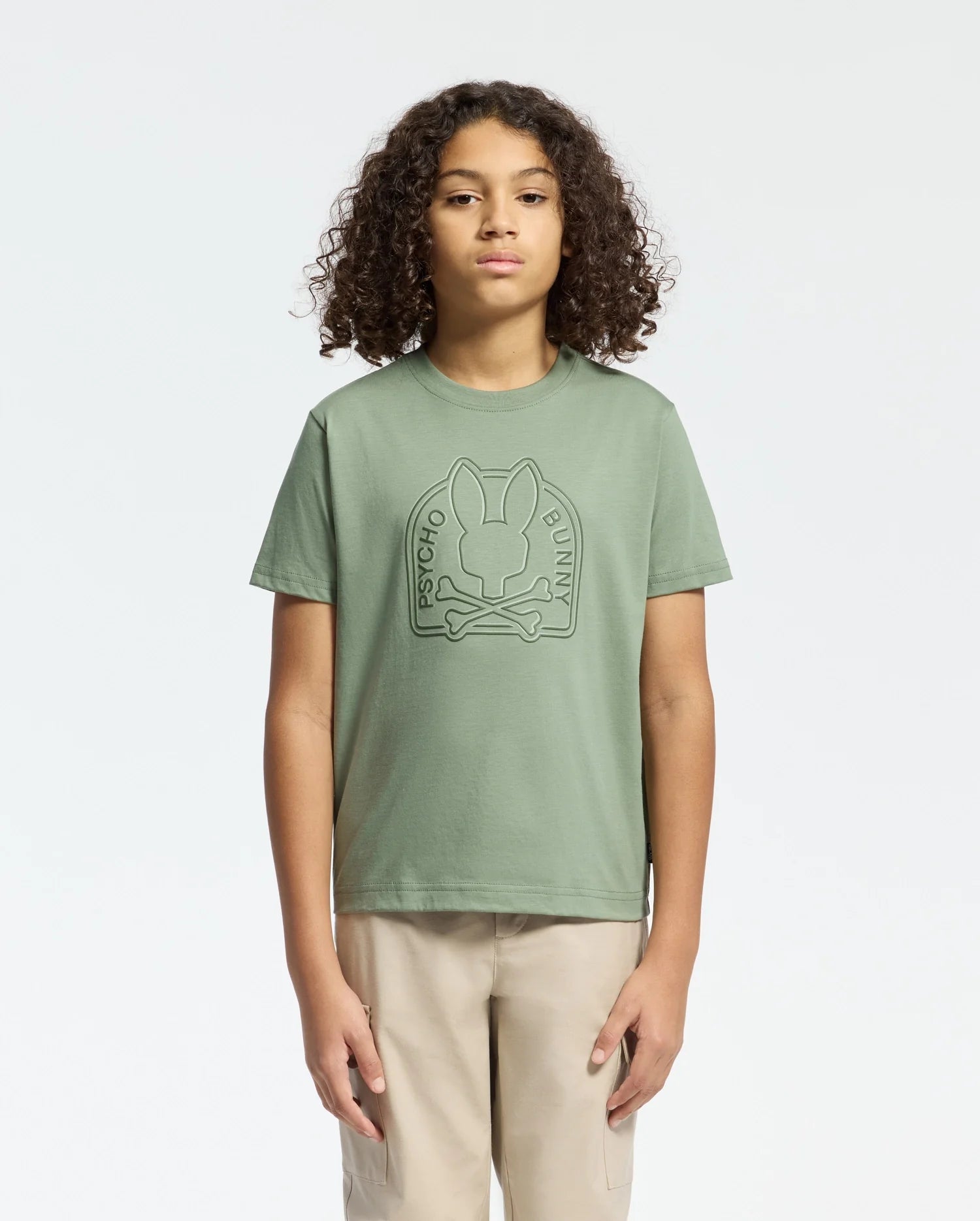 With curly hair and beige pants, a person faces forward wearing the KIDS MELBOURNE GRAPHIC TEE (B0U410F200) by Psycho Bunny, featuring a bold Bunny graphic on green Pima cotton, set against a plain white background.