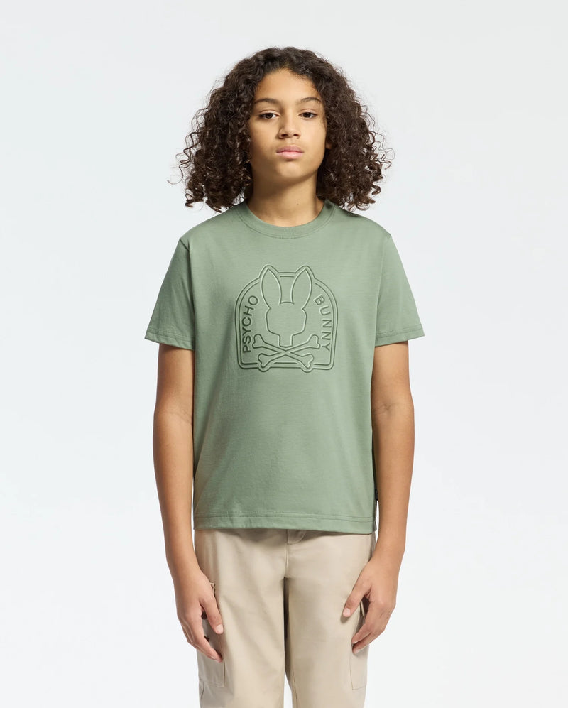 With curly hair and beige pants, a person faces forward wearing the KIDS MELBOURNE GRAPHIC TEE (B0U410F200) by Psycho Bunny, featuring a bold Bunny graphic on green Pima cotton, set against a plain white background.
