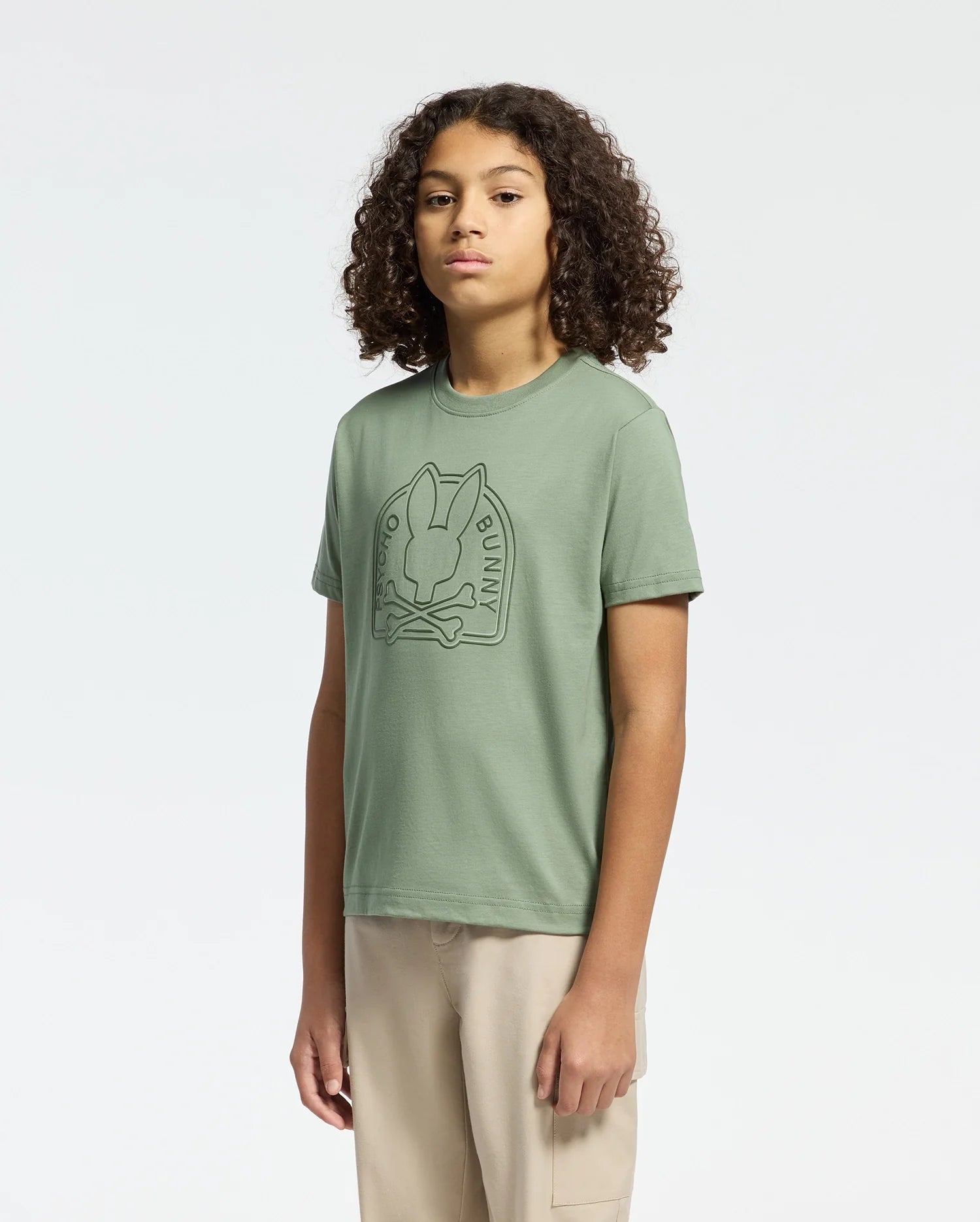 A person with curly hair stands against a plain background, wearing a green KIDS MELBOURNE GRAPHIC TEE (B0U410F200) by Psycho Bunny and beige pants.