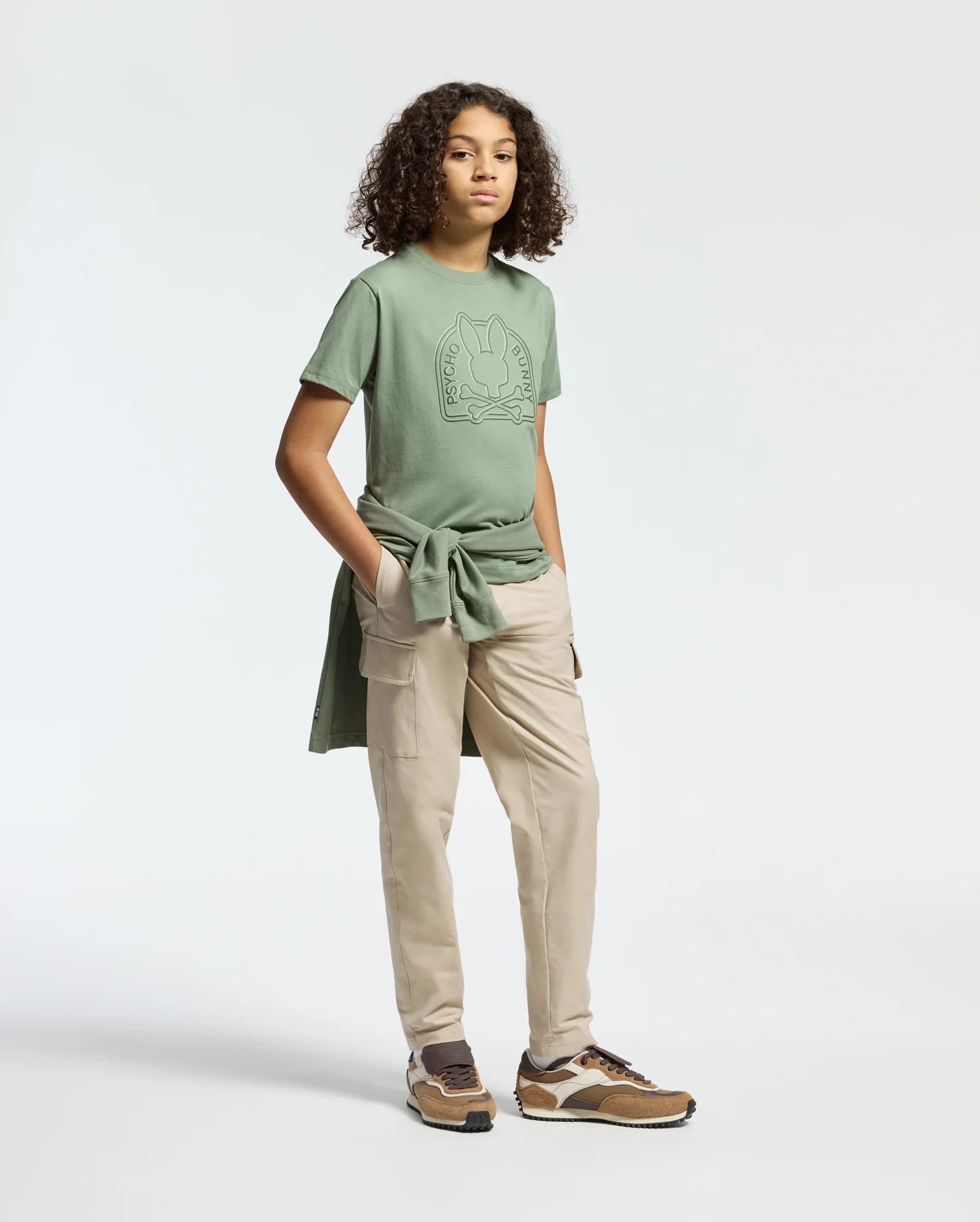 In a simple studio, a curly-haired person wears a casual menKnit outfit with the Psycho Bunny KIDS MELBOURNE GRAPHIC TEE in green, light khaki cargo pants, brown sneakers, and ties a Pima cotton green sweatshirt around their waist.
