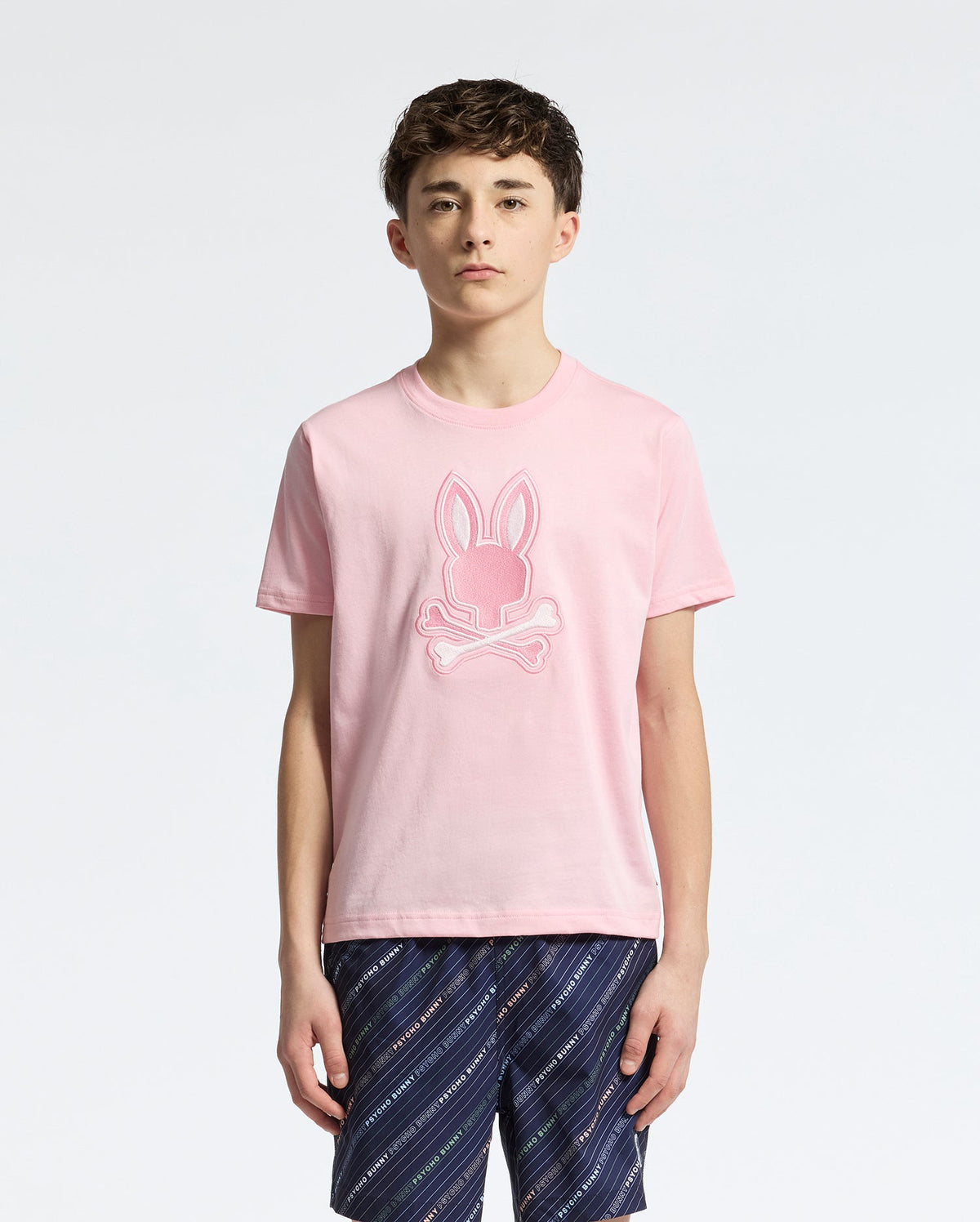 A young person, with short brown hair and dark patterned shorts, stands facing forward in a Psycho Bunny KIDS NEW CASTLE EMBROIDERED GRAPHIC TEE - B0U415F200. The T-shirt is premium Pima cotton, pink, and features an embroidered bunny skull design against a plain white background.