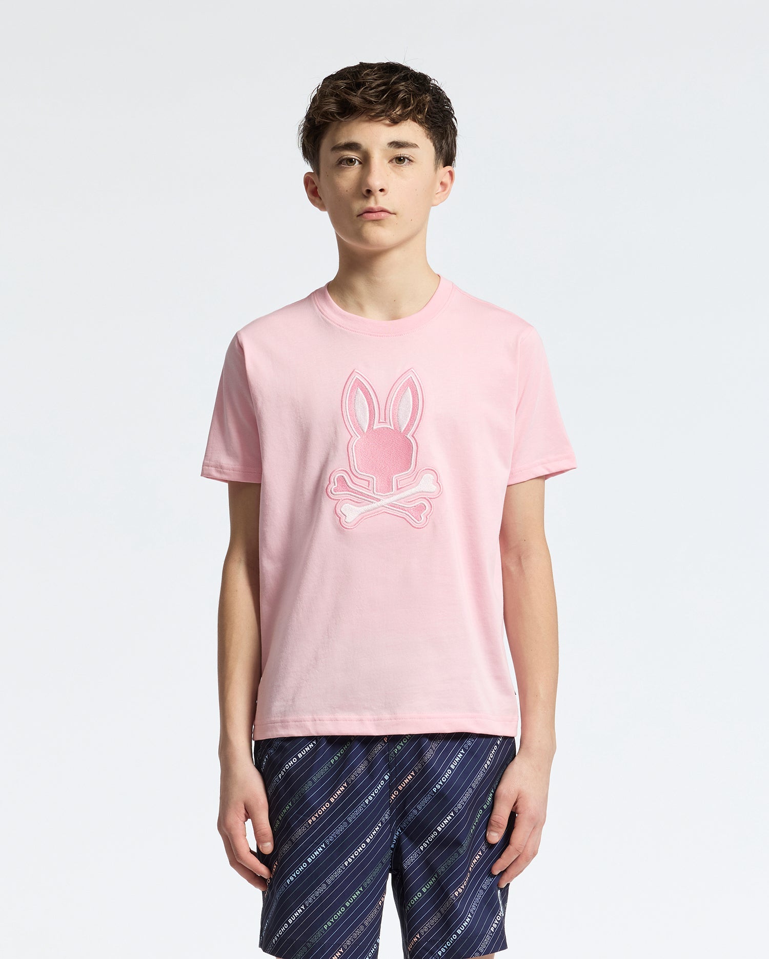 A young person, with short brown hair and dark patterned shorts, stands facing forward in a Psycho Bunny KIDS NEW CASTLE EMBROIDERED GRAPHIC TEE - B0U415F200. The T-shirt is premium Pima cotton, pink, and features an embroidered bunny skull design against a plain white background.