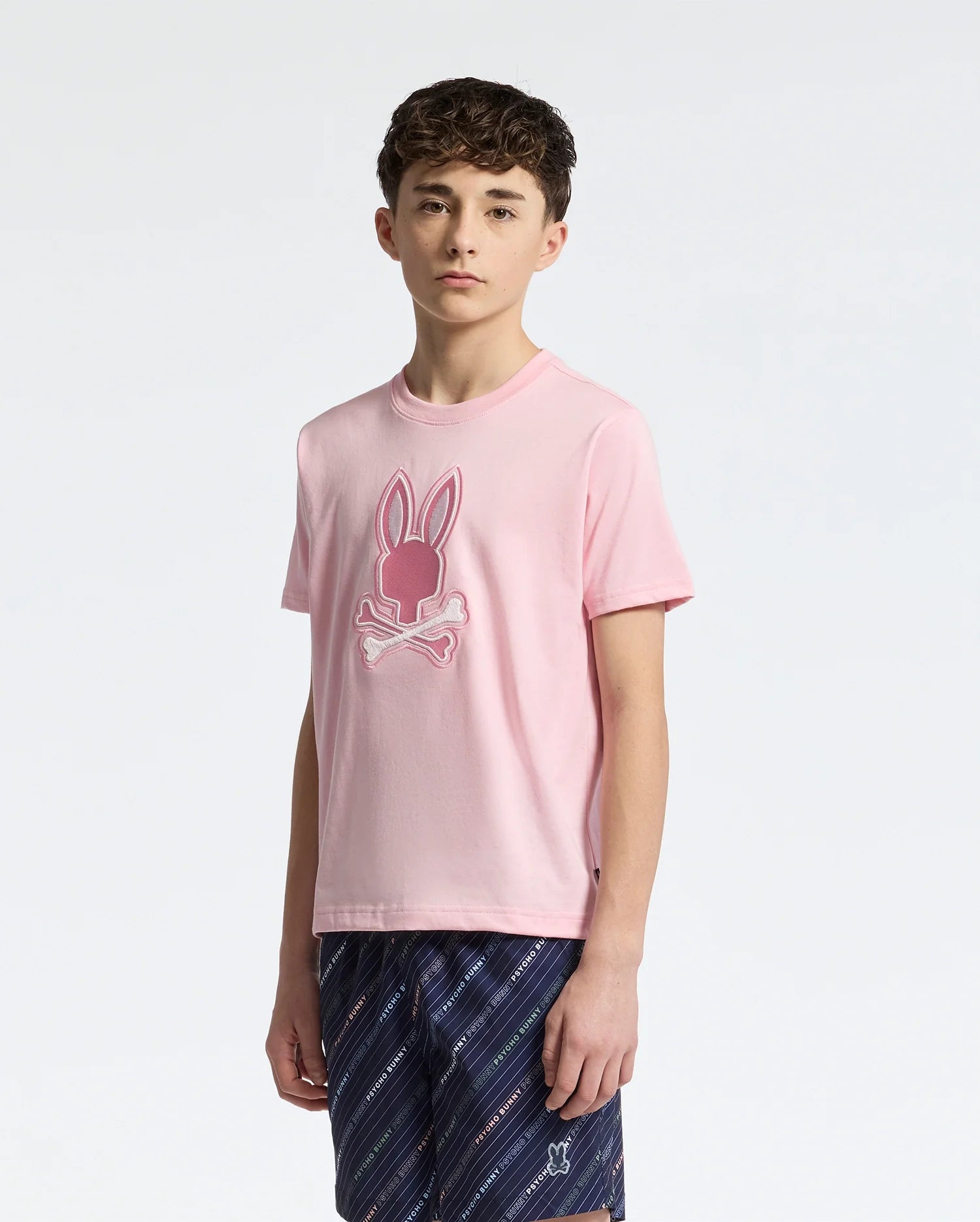 A teen sports the Psycho Bunny KIDS NEW CASTLE EMBROIDERED GRAPHIC TEE in pink, made from premium Pima cotton, showcasing a Bunny skull and crossbones design. They pair it with blue plaid shorts against a plain white backdrop.