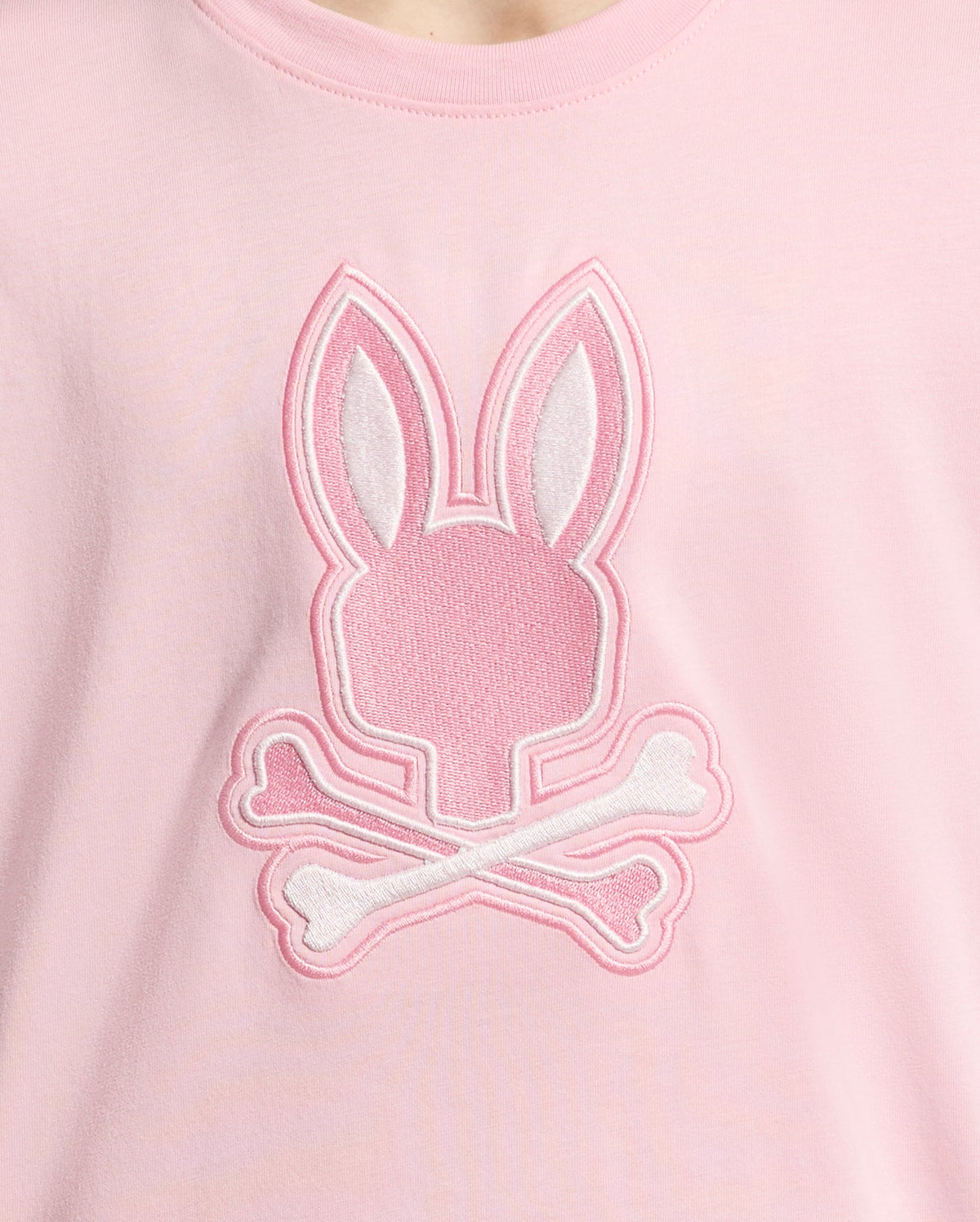 The KIDS NEW CASTLE EMBROIDERED GRAPHIC TEE by Psycho Bunny is a pink shirt made from premium Pima cotton, featuring an embroidered Bunny logo with a stylized head and long ears over two crossed bones, resembling a fun skull-and-crossbones graphic.