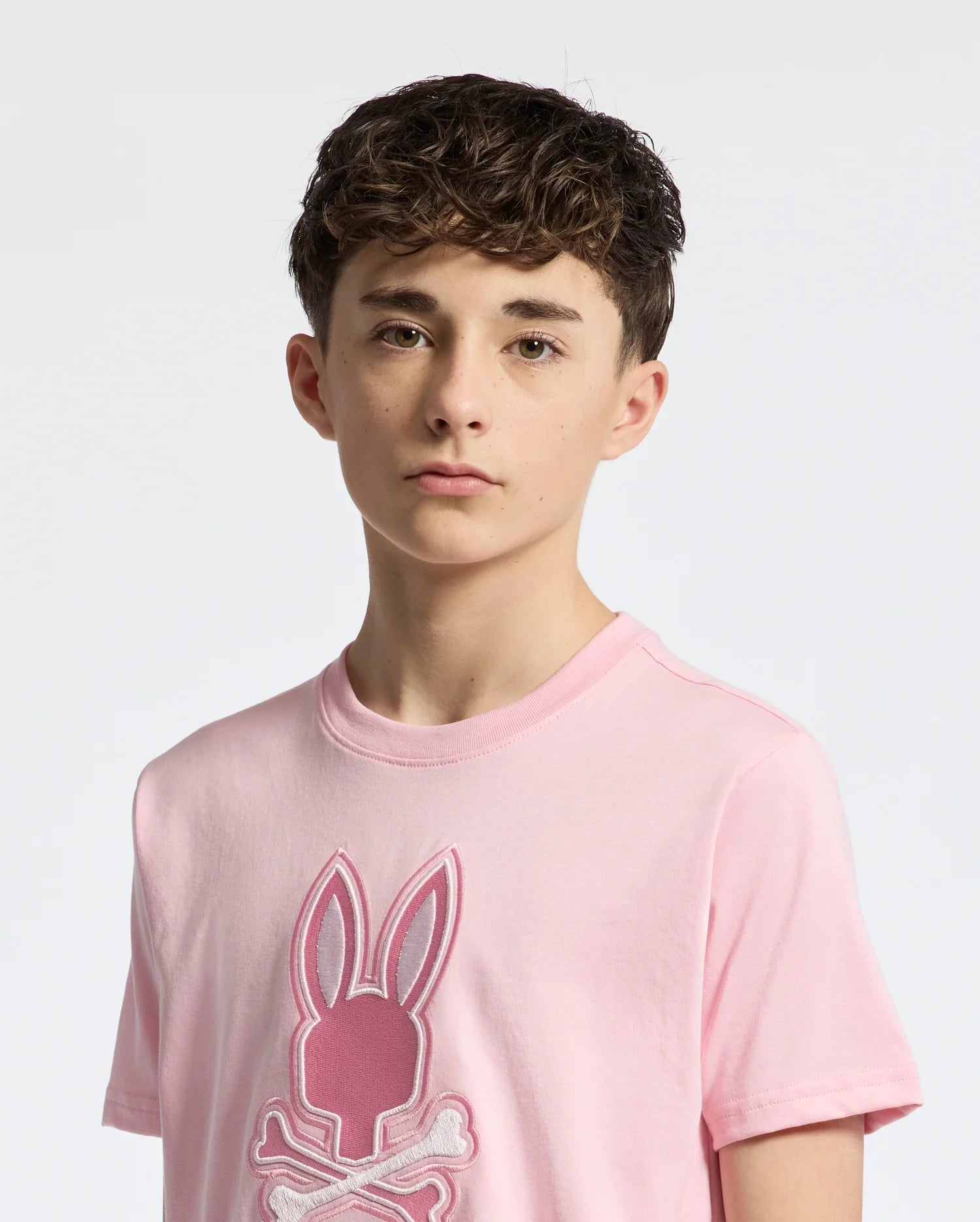 A young person with short, curly brown hair wears a light pink Psycho Bunny KIDS NEW CASTLE EMBROIDERED GRAPHIC TEE featuring a stylized bunny and crossbones logo, made from premium Pima cotton. They have a neutral expression against a plain light background.
