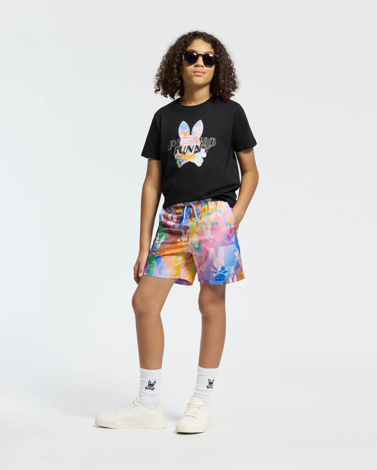 A person with curly hair wearing sunglasses and a Psycho Bunny KIDS MILOS GRAPHIC TEE in black confidently dons vibrant multicolored shorts, white socks, and sleek white sneakers.