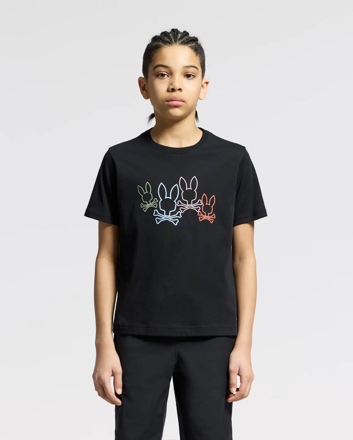 A person wears the Psycho Bunny KIDS SILVERDALE GRAPHIC TEE, featuring a vibrant bunny graphic with colorful outlines of three rabbits on soft Pima cotton. They stand against a plain background, their braided hair and neutral expression adding understated elegance.