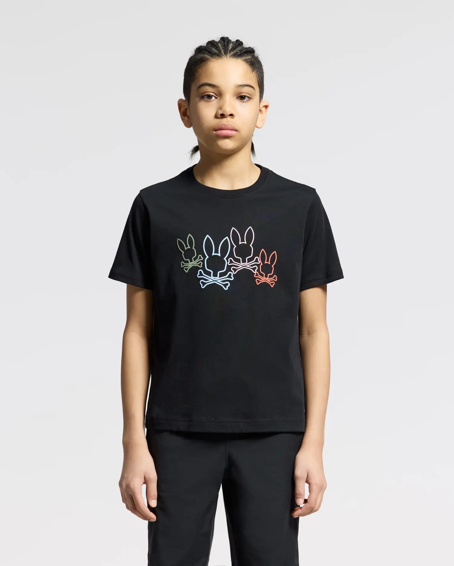 A person wears the Psycho Bunny KIDS SILVERDALE GRAPHIC TEE, featuring a vibrant bunny graphic with colorful outlines of three rabbits on soft Pima cotton. They stand against a plain background, their braided hair and neutral expression adding understated elegance.