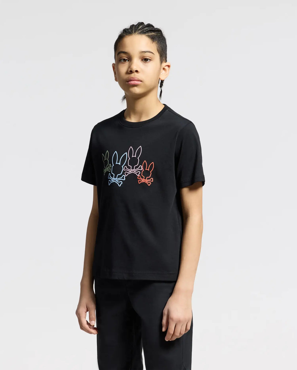 A person with braided hair stands against a plain background, wearing the KIDS SILVERDALE GRAPHIC TEE - B0U499F200 by Psycho Bunny. The black tee features colorful abstract bunny graphics and is crafted from soft Pima cotton, perfectly complementing sleek black pants.