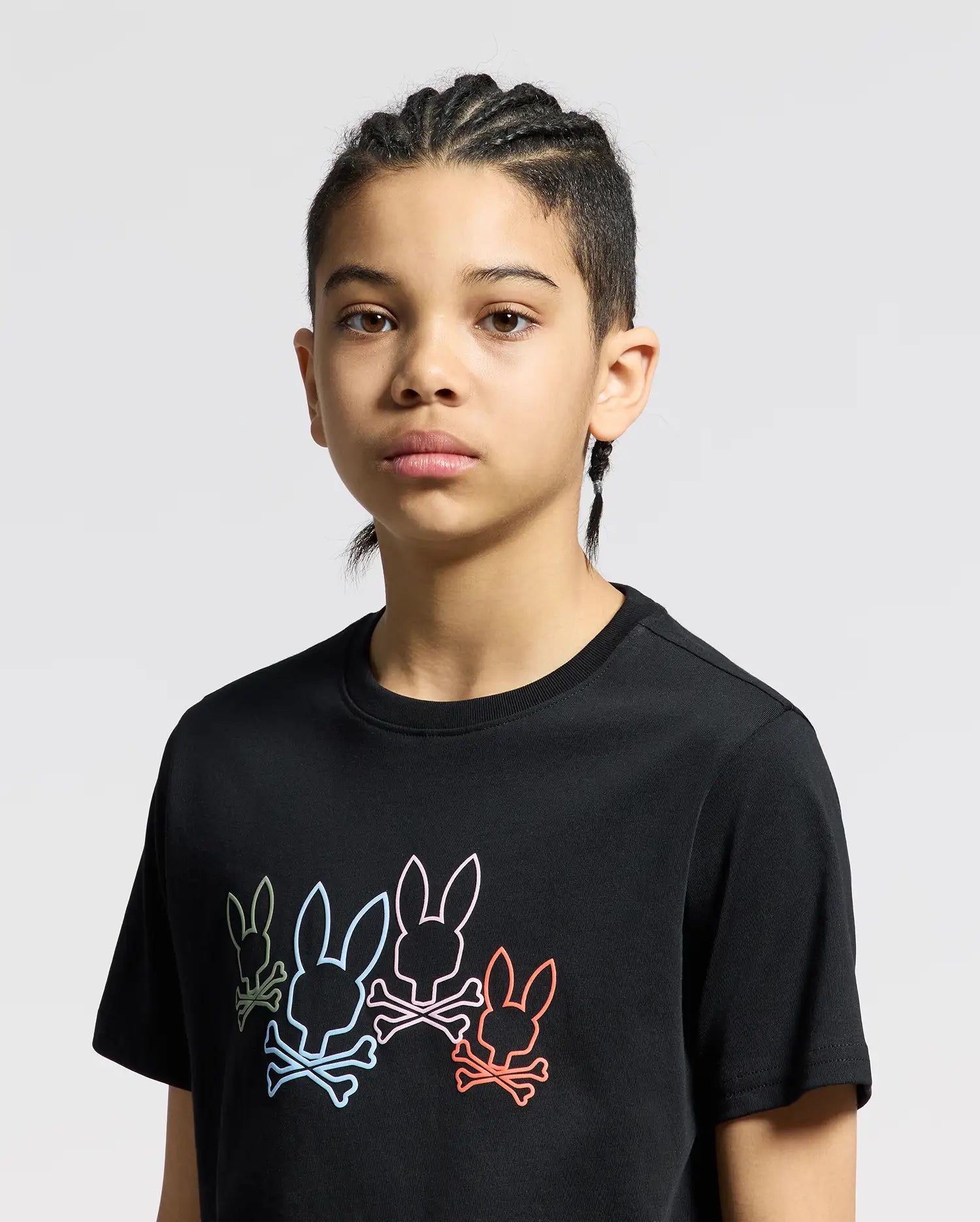 A young person with braided hair wears the KIDS SILVERDALE GRAPHIC TEE by Psycho Bunny, a black Pima cotton shirt showcasing colorful bunny graphics across the chest. The water-based print stands out against the light gray background.