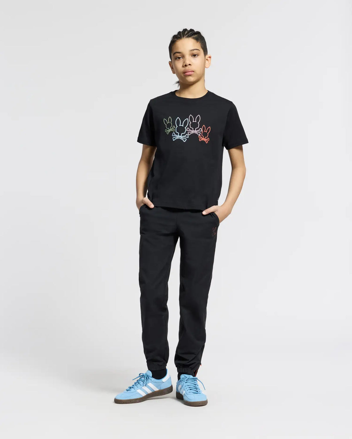 A young person poses confidently against a plain background, hands in pockets. They're wearing Psycho Bunny's KIDS SILVERDALE GRAPHIC TEE (B0U499F200), black pants, and blue sneakers, featuring colorful bunny graphics on the black Pima cotton top while looking at the camera.