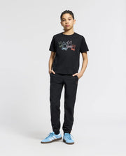 A young person poses confidently against a plain background, hands in pockets. They're wearing Psycho Bunny's KIDS SILVERDALE GRAPHIC TEE (B0U499F200), black pants, and blue sneakers, featuring colorful bunny graphics on the black Pima cotton top while looking at the camera.