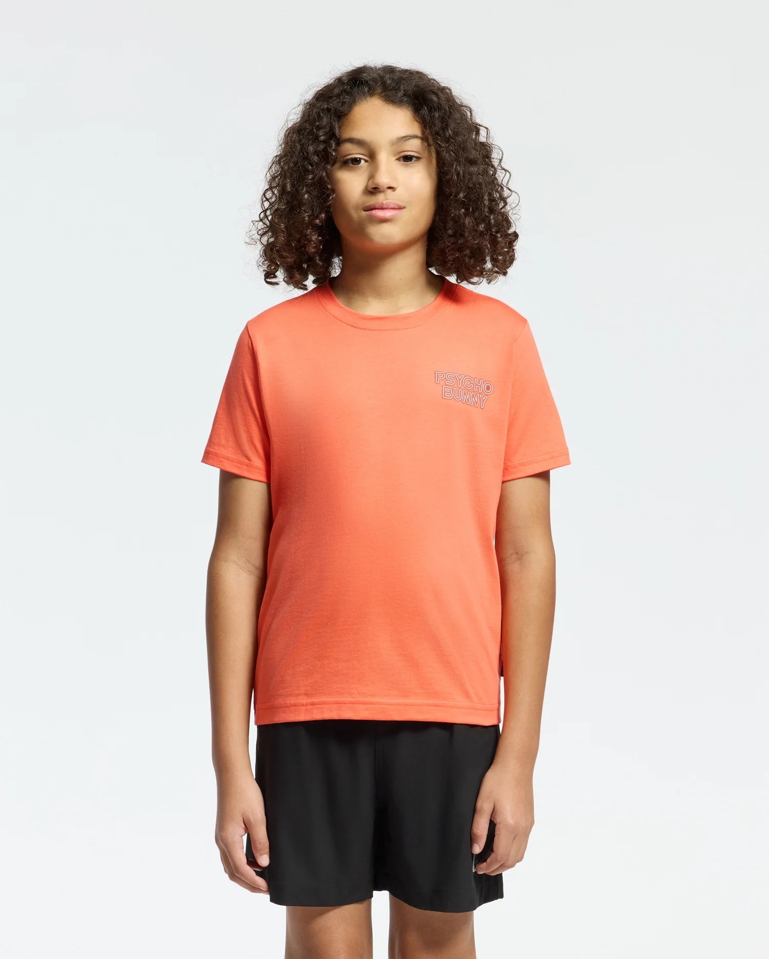 A child with curly hair faces forward wearing a regular fit, bright orange Psycho Bunny KIDS SYDNEY BACK GRAPHIC TEE (B0U509F200) and black shorts against a plain white background.