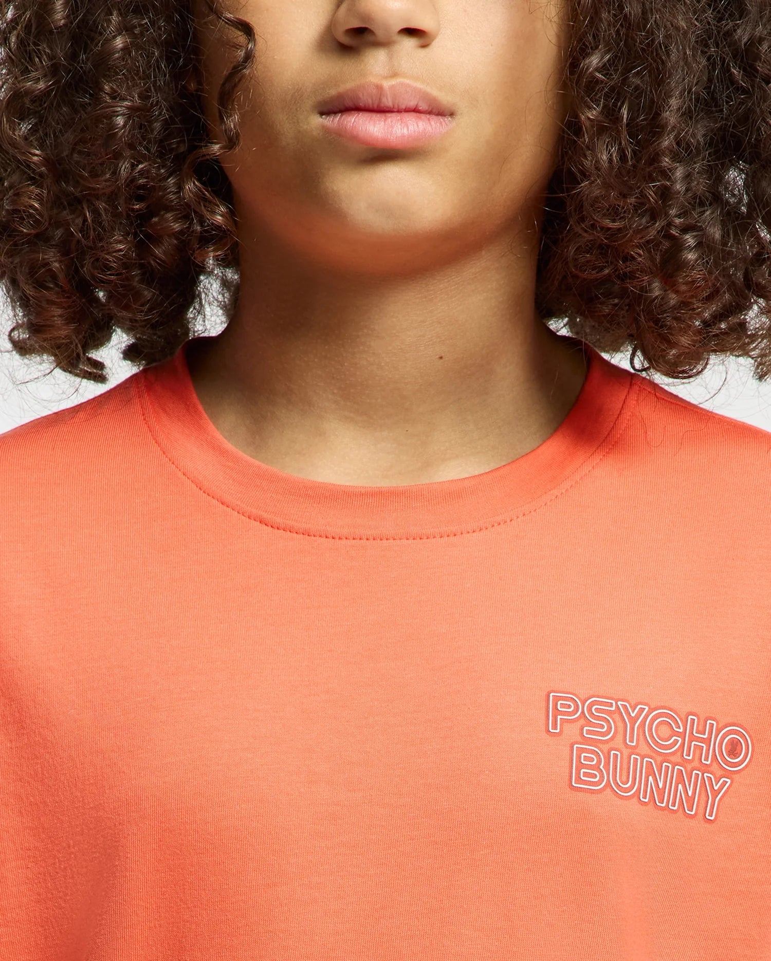 A child with curly hair wears the Psycho Bunny KIDS SYDNEY BACK GRAPHIC TEE in orange, featuring a water-based "PSYCHO BUNNY" print on the left chest. The photo is cropped to show only the lower face and upper torso.