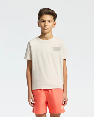 A young person with short hair and a neutral expression stands against a plain background wearing the light beige Psycho Bunny KIDS SYDNEY BACK GRAPHIC TEE, B0U509F200, in Pima cotton with a subtle left chest water-based print, paired with coral shorts.