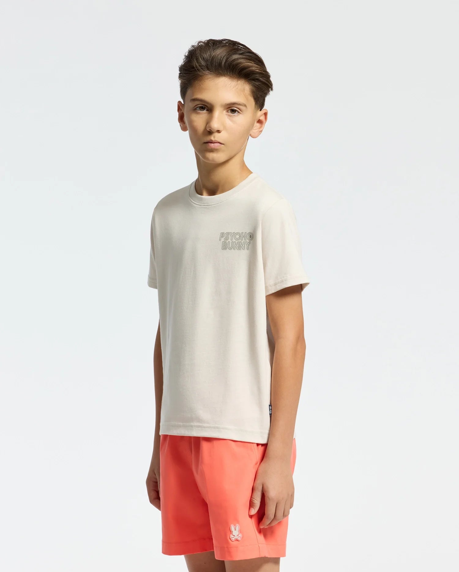 A young individual confidently wears the Psycho Bunny KIDS SYDNEY BACK GRAPHIC TEE - B0U509F200, a soft Pima cotton, light-toned shirt, paired with bright coral shorts. In a regular fit style, they stand against a plain background with hands at their sides, looking directly at the camera.
