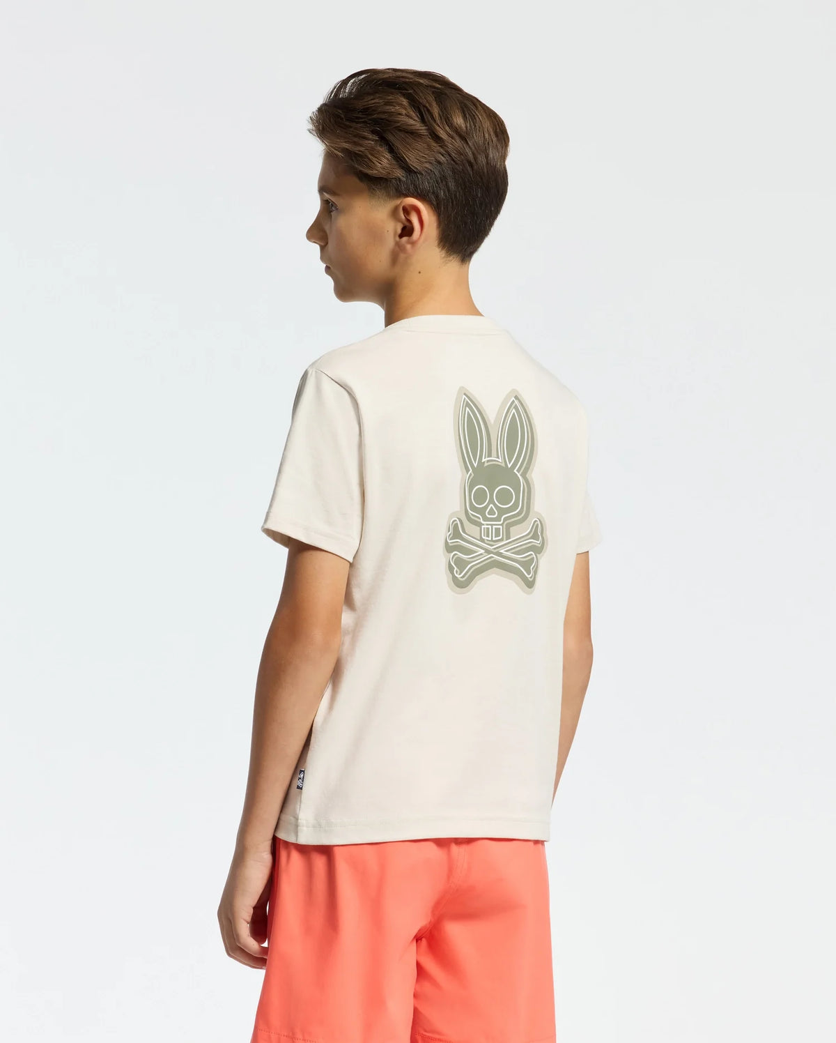 A young boy stands slightly sideways against a plain white background, wearing the KIDS SYDNEY BACK GRAPHIC TEE by Psycho Bunny, a beige Pima cotton T-shirt featuring a large rabbit skull and crossbones graphic in water-based print on the back. He pairs it with coral-colored shorts.