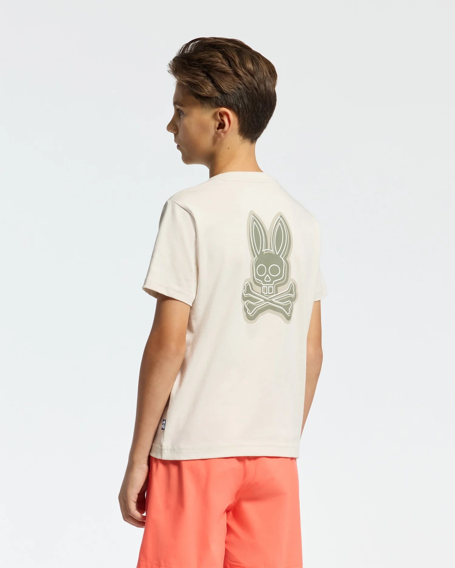 A young boy stands slightly sideways against a plain white background, wearing the KIDS SYDNEY BACK GRAPHIC TEE by Psycho Bunny, a beige Pima cotton T-shirt featuring a large rabbit skull and crossbones graphic in water-based print on the back. He pairs it with coral-colored shorts.