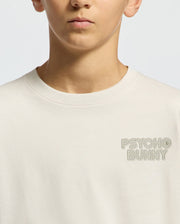 A teen in a light beige KIDS SYDNEY BACK GRAPHIC TEE by Psycho Bunny, tailored in regular fit Pima cotton, features subtle "PSYCHO BUNNY" embroidery on the chest against a plain light gray background.