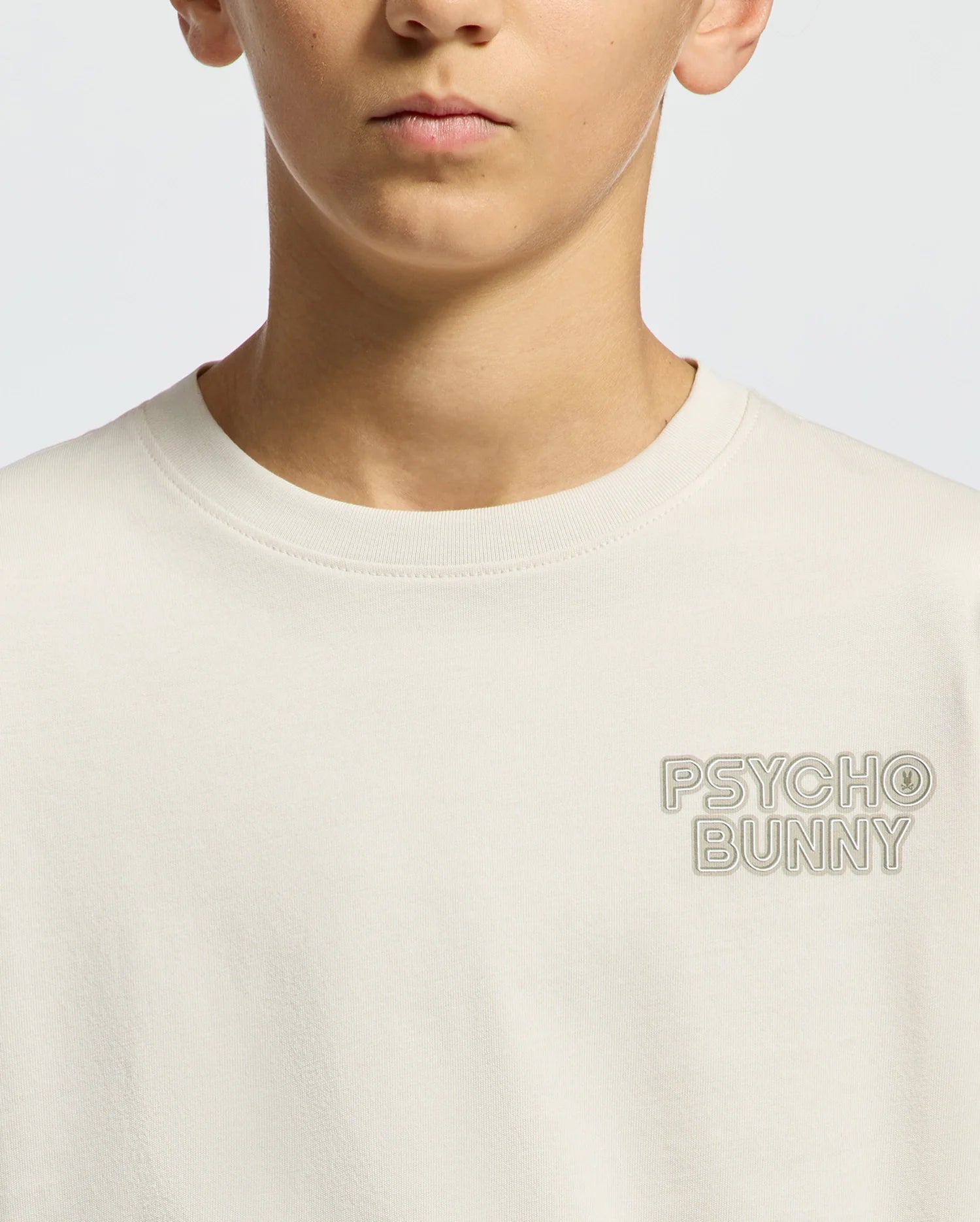 A teen in a light beige KIDS SYDNEY BACK GRAPHIC TEE by Psycho Bunny, tailored in regular fit Pima cotton, features subtle "PSYCHO BUNNY" embroidery on the chest against a plain light gray background.