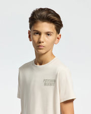 A boy with short brown hair wears a light-colored KIDS SYDNEY BACK GRAPHIC TEE by Psycho Bunny, featuring "Psycho Bunny" text in a stylish water-based print on the chest. He stands against a plain background, looking directly at the camera.
