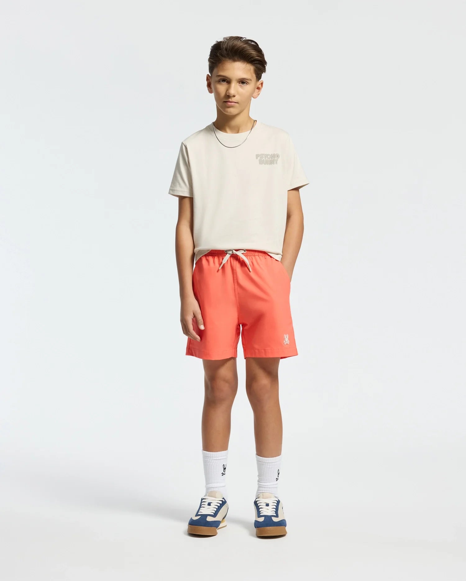 A young person stands against a plain background, wearing the Psycho Bunny KIDS SYDNEY BACK GRAPHIC TEE in light beige, bright coral shorts, and white socks with patterned emblems. Their sneakers complete the regular fit look as they rest hands in pockets, their short hair styled perfectly.