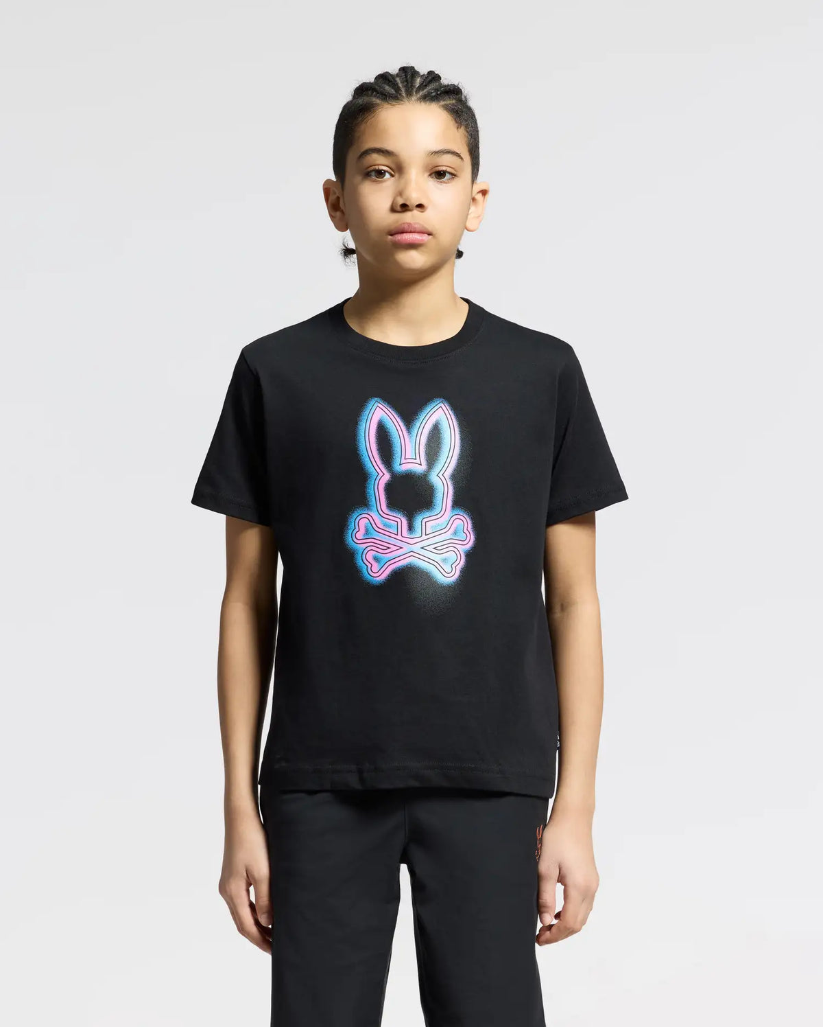 A young person with braided hair stands calmly against a neutral background, wearing the Psycho Bunny KIDS ANDROS GRAPHIC TEE in black. The luxurious Pima Cotton shirt features a neon bunny outline with crossed bones underneath.
