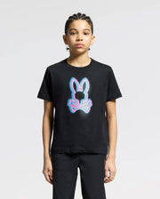 A young person with braided hair stands calmly against a neutral background, wearing the Psycho Bunny KIDS ANDROS GRAPHIC TEE in black. The luxurious Pima Cotton shirt features a neon bunny outline with crossed bones underneath.