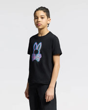 A young individual with braided hair models a Psycho Bunny KIDS ANDROS GRAPHIC TEE (B0U525F200) in black, crafted from Pima Cotton in Peru. It showcases a neon bunny with crossed bones design, set against a plain light grey backdrop.
