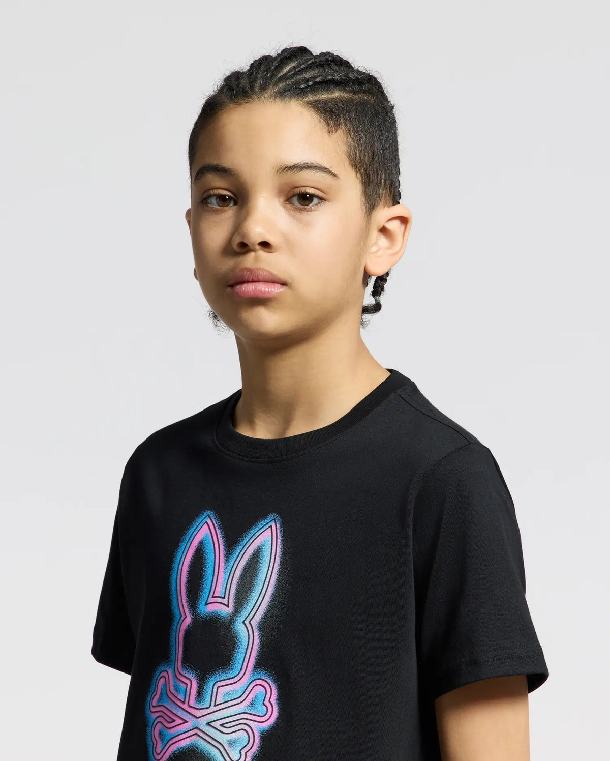 A young person with cornrows wears a Kids Andros Graphic Tee in black by Psycho Bunny. Made in Peru from soft Pima Cotton, it features a neon-style bunny and crossbones graphic. They stand neutrally against a plain, light background.