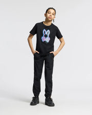 A young person stands confidently in the KIDS ANDROS GRAPHIC TEE - B0U525F200 from Psycho Bunny, a black Pima cotton shirt featuring a neon bunny design. With hands in the pockets of black pants and wearing black sneakers, they pose against a plain white background.