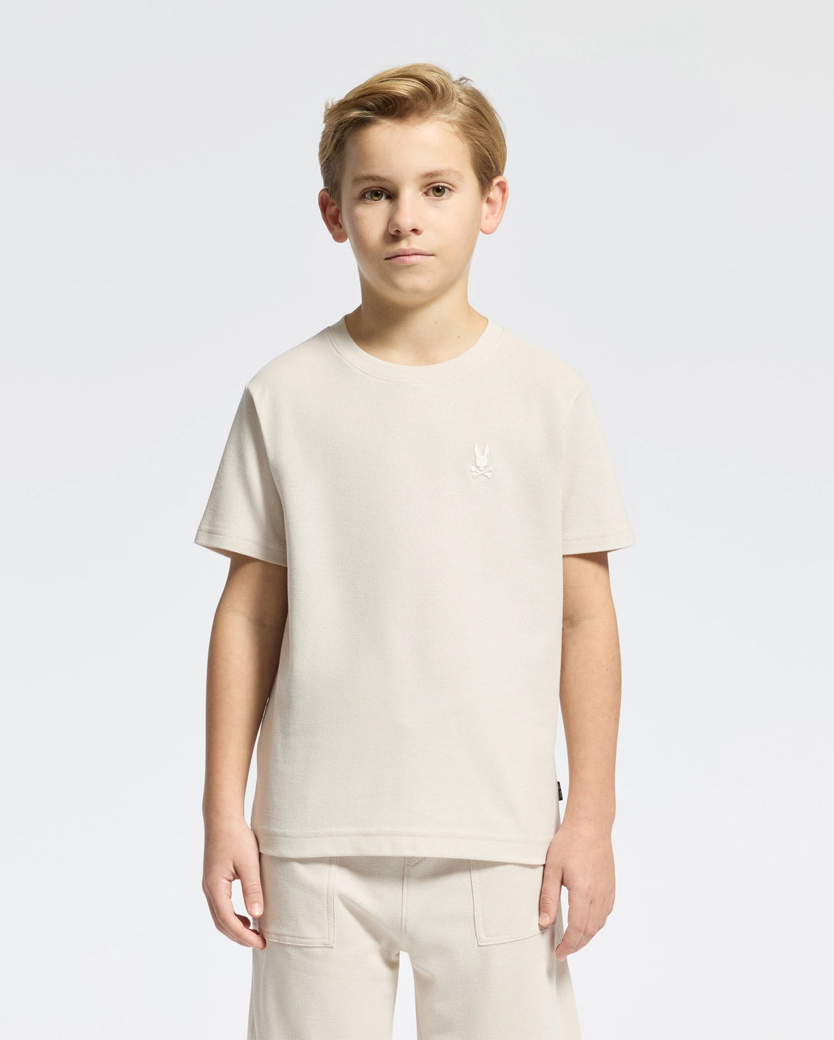 A young boy with short hair models a classic style Psycho Bunny KIDS TAYLOR PIQUE TEE - B0U579F200 in light beige Pima cotton alongside matching pants on a plain white background. The tee showcases an embroidered Bunny emblem on the chest as he gazes at the camera neutrally.