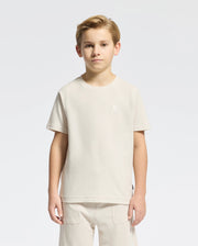 A young boy with short hair models a classic style Psycho Bunny KIDS TAYLOR PIQUE TEE - B0U579F200 in light beige Pima cotton alongside matching pants on a plain white background. The tee showcases an embroidered Bunny emblem on the chest as he gazes at the camera neutrally.