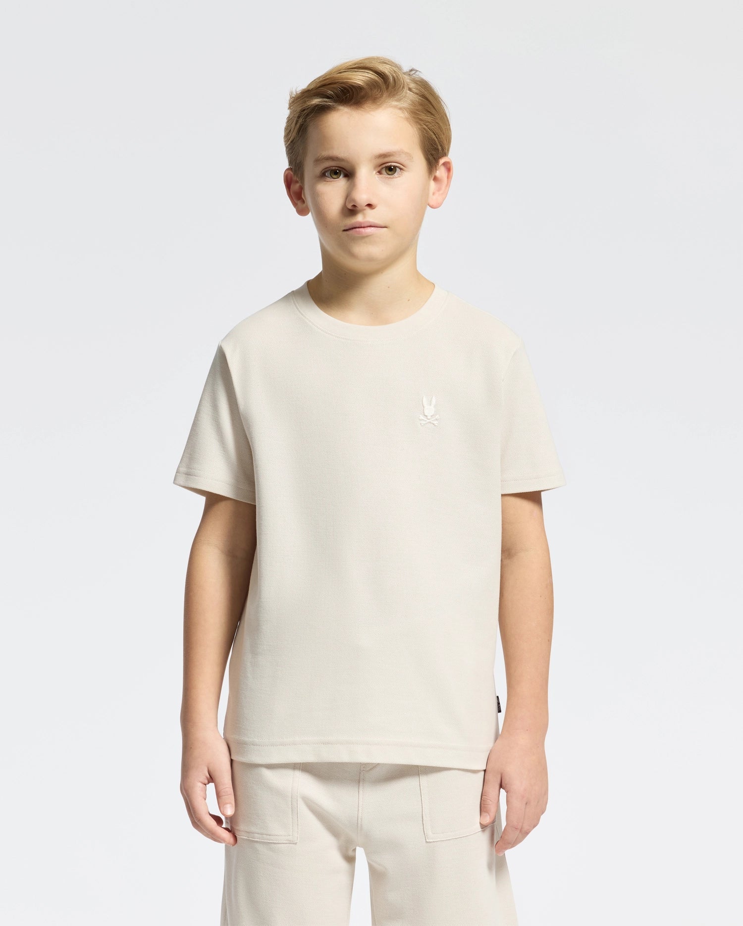 A young boy with short hair models a classic style Psycho Bunny KIDS TAYLOR PIQUE TEE - B0U579F200 in light beige Pima cotton alongside matching pants on a plain white background. The tee showcases an embroidered Bunny emblem on the chest as he gazes at the camera neutrally.