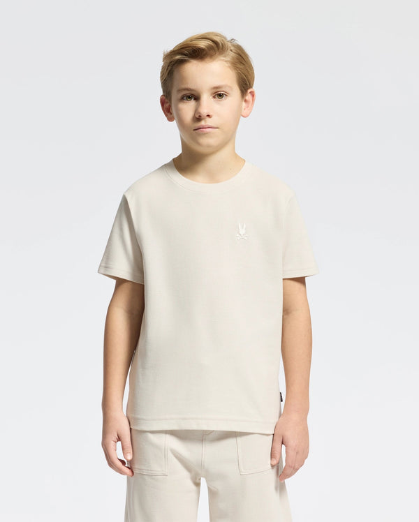 A young boy with short hair models a classic style Psycho Bunny KIDS TAYLOR PIQUE TEE - B0U579F200 in light beige Pima cotton alongside matching pants on a plain white background. The tee showcases an embroidered Bunny emblem on the chest as he gazes at the camera neutrally.