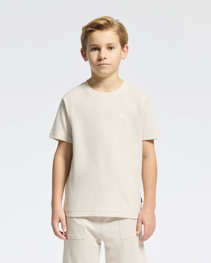 A young boy with short hair models a classic style Psycho Bunny KIDS TAYLOR PIQUE TEE - B0U579F200 in light beige Pima cotton alongside matching pants on a plain white background. The tee showcases an embroidered Bunny emblem on the chest as he gazes at the camera neutrally.