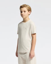 A young blond boy stands against a white background, wearing the beige Psycho Bunny KIDS TAYLOR PIQUE TEE (B0U579F200) and matching Pima cotton pants. An embroidered bunny adds charm to his outfit as he looks at the camera with a neutral expression.