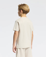A young boy in the KIDS TAYLOR PIQUE TEE by Psycho Bunny, with short blond hair, stands facing away. He wears matching beige shorts crafted from soft Pima cotton. The classic style features an embroidered bunny on a white background.