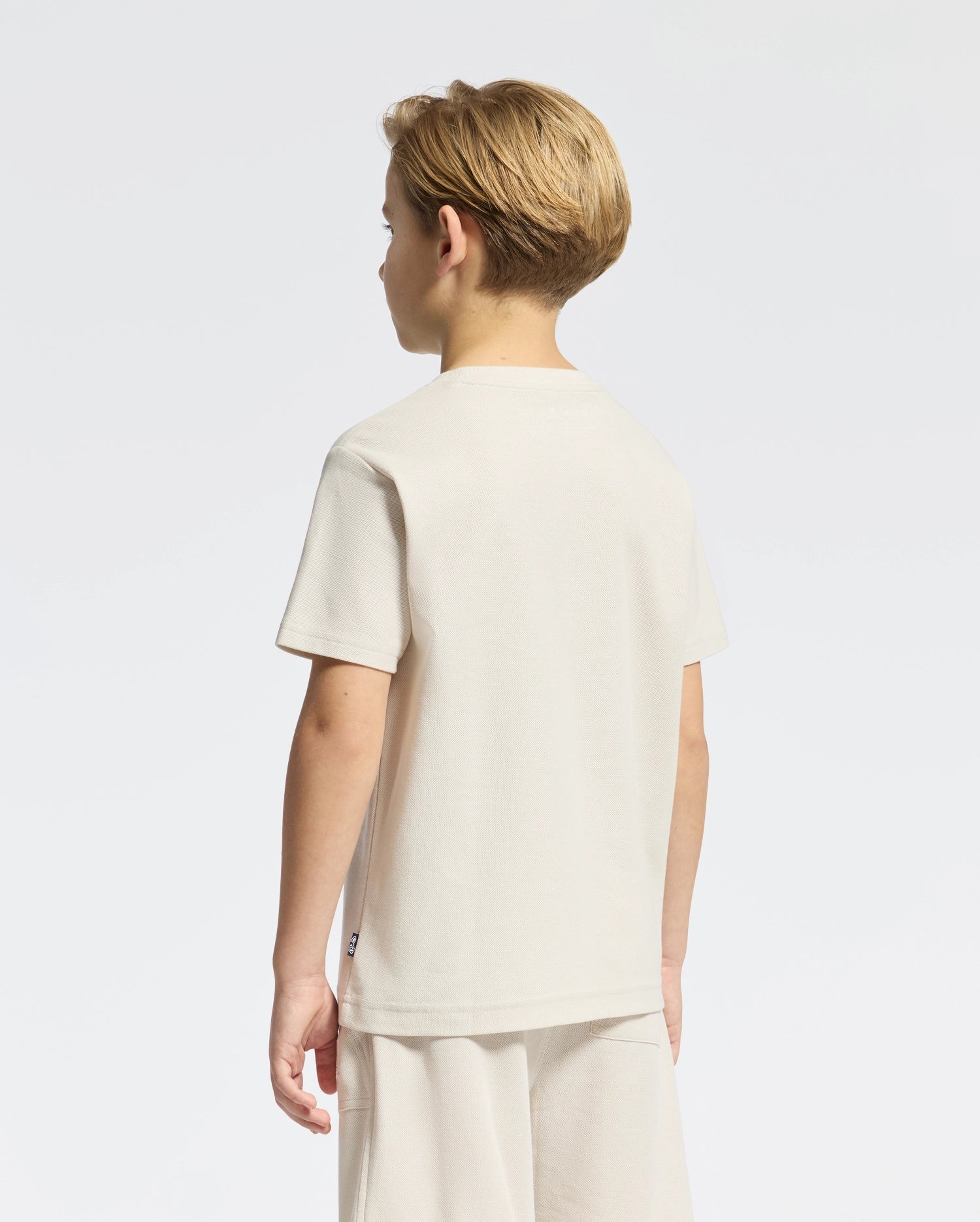 A young boy in the KIDS TAYLOR PIQUE TEE by Psycho Bunny, with short blond hair, stands facing away. He wears matching beige shorts crafted from soft Pima cotton. The classic style features an embroidered bunny on a white background.