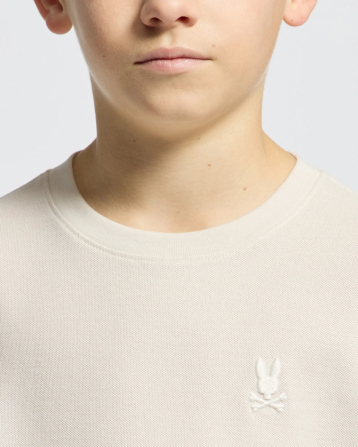 A person is seen wearing a light beige Pima cotton sweater with a textured design and the Psycho Bunny emblem, featuring rabbit ears and crossbones, on the chest. The image highlights the sweater's classic style against a simple backdrop, focusing on their neck and lower face.