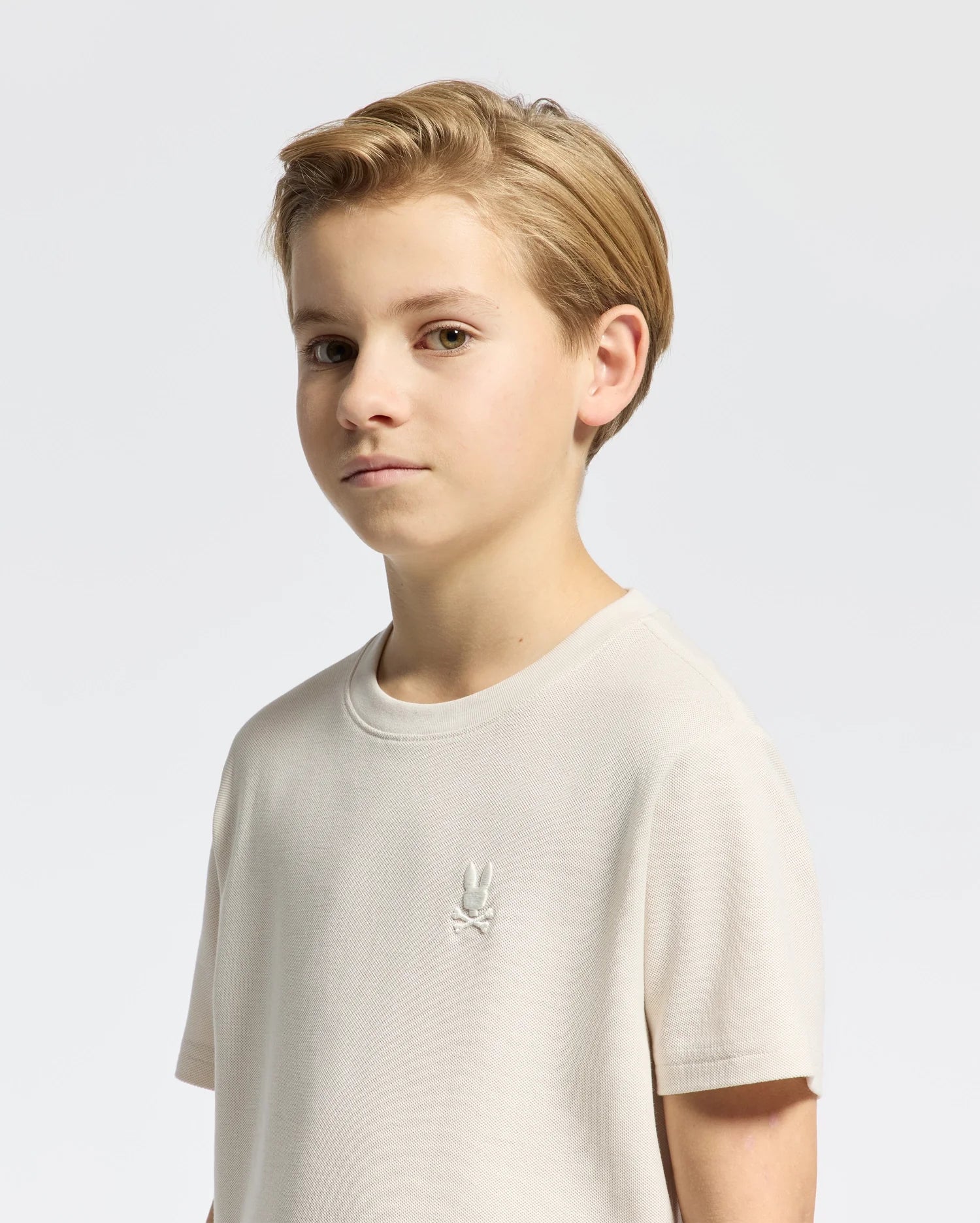 A young person with short, blonde hair stands against a plain background, wearing the KIDS TAYLOR PIQUE TEE by Psycho Bunny. This light-colored T-shirt, made from soft Pima cotton, features an embroidered bunny emblem above crossed bones on the chest for a classic touch.