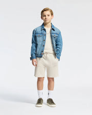 A young boy radiates classic style in a blue denim jacket, light beige Pima cotton shirt, and drawstring shorts. His look is completed with white socks, olive green sneakers, and a subtle embroidered logo from Psycho Bunny's KIDS TAYLOR PIQUE TEE as he stands against a light gray background.