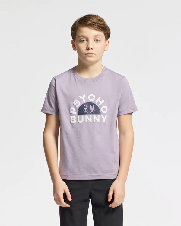 A young person with short, light brown hair stands against a white background wearing the KIDS GOLDIE GRAPHIC TEE in lavender by Psycho Bunny, made from luxurious Peruvian Pima cotton. The tee features "Psycho Bunny" and a playful bunny graphic in white text, paired with sleek black pants.