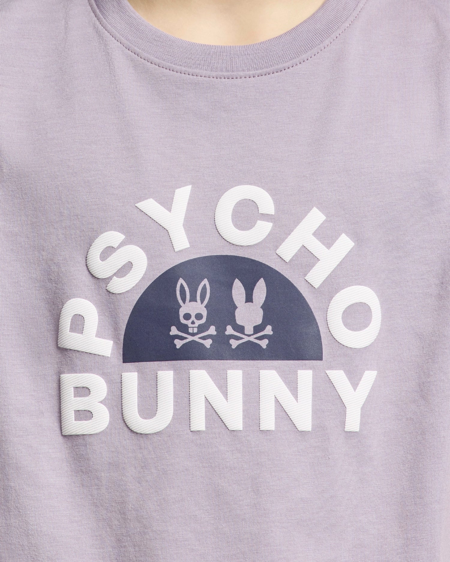 The KIDS GOLDIE GRAPHIC TEE - B0U606F200 from Psycho Bunny, made of luxurious Peruvian Pima cotton, features "Psycho Bunny" in white around a half-circle with two stylized bunnies and crossed bones forming a striking graphic on a lavender backdrop.