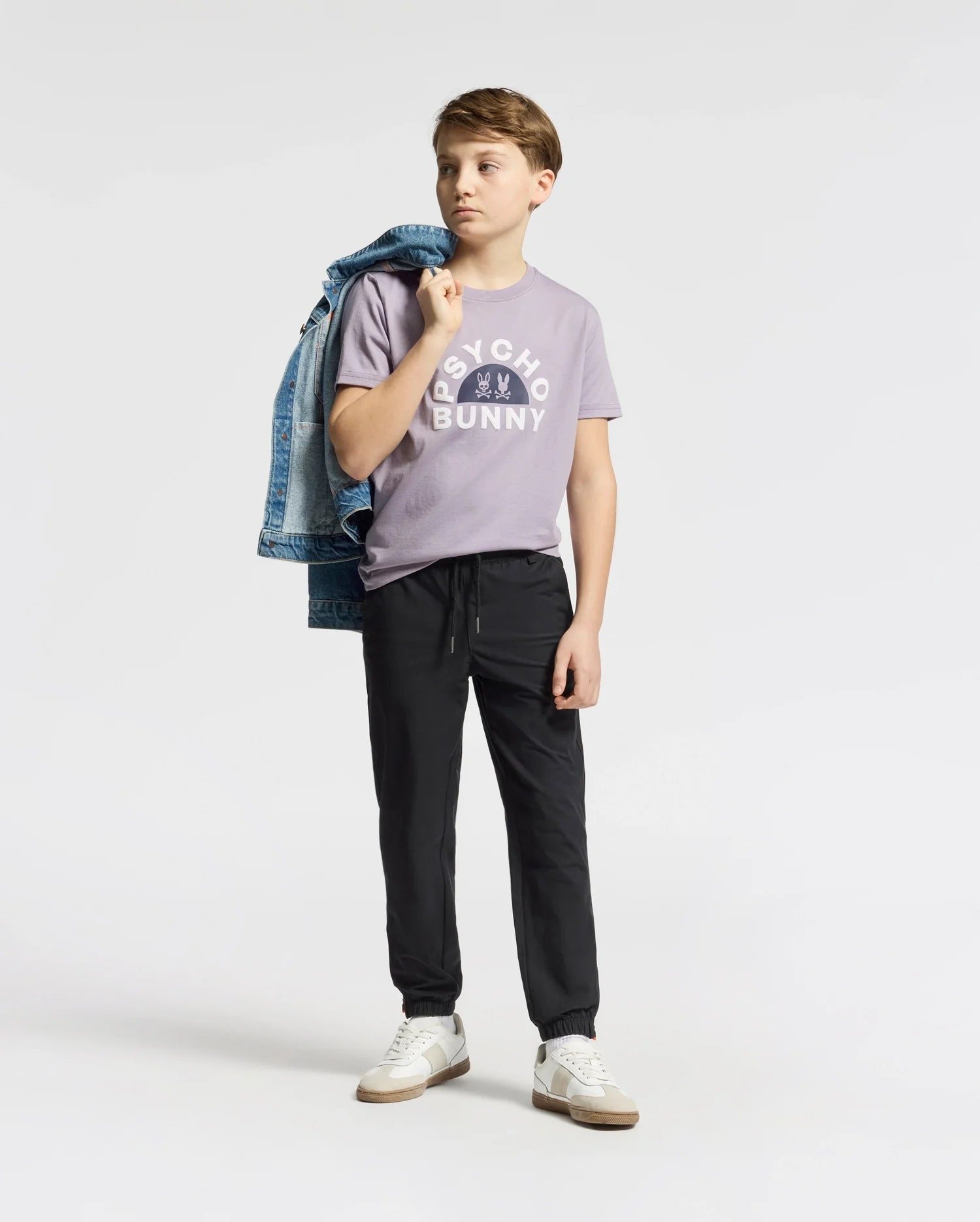 A young person stands against a plain background wearing a purple KIDS GOLDIE GRAPHIC TEE by Psycho Bunny, black pants, and white sneakers. They drape a blue denim jacket over one shoulder and gaze thoughtfully to the side.