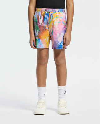 A person sports Psycho Bunny's vibrant KIDS MILOS PRINTED SWIM TRUNK (B0W446F200) with abstract patterns, paired with classic white socks and sneakers. The light backdrop highlights the laid-back, quick-dry fabric of the ensemble.