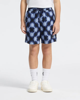 A person wearing KIDS PAROS PRINTED SWIM TRUNK by Psycho Bunny, a white shirt, white socks, and sneakers stands against a plain backdrop. The blue and white abstract design on the swim trunks pops alongside the logo on the socks for a crisp look.